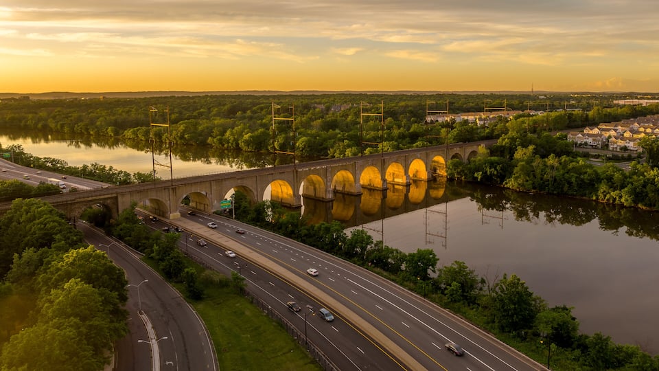 A bridge with multiple arches over a river, surrounded by greenery and a highway.