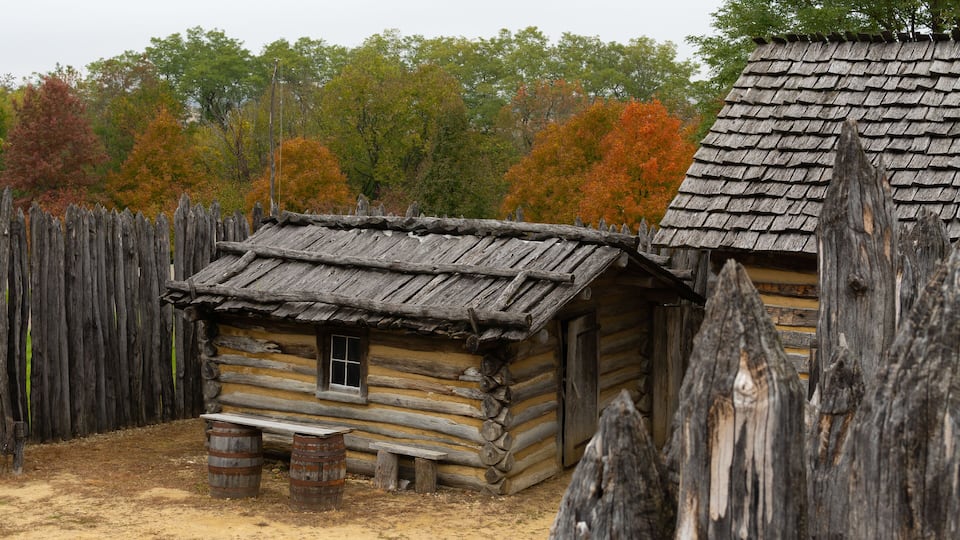 A rustic log cabin with a thatched roof, surrounded by a wooden fence and autumn-colored trees.