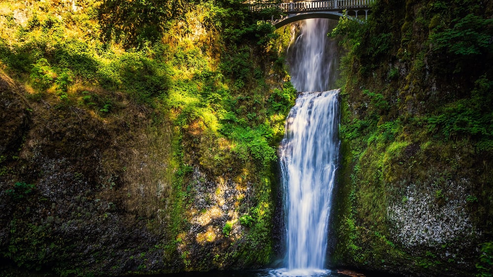 Ein Wasserfall stürzt eine von Moos bewachsene Klippe hinab, eine Brücke überspannt die Spitze.