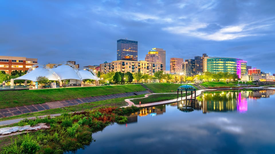A city skyline at dusk with a calm river reflecting the buildings, a park with a white gazebo, and a modern building with a blue dome.