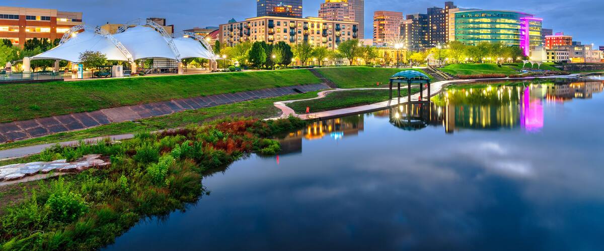 A city skyline at dusk with a calm river reflecting the buildings, a park with a white gazebo, and a modern building with a blue dome.