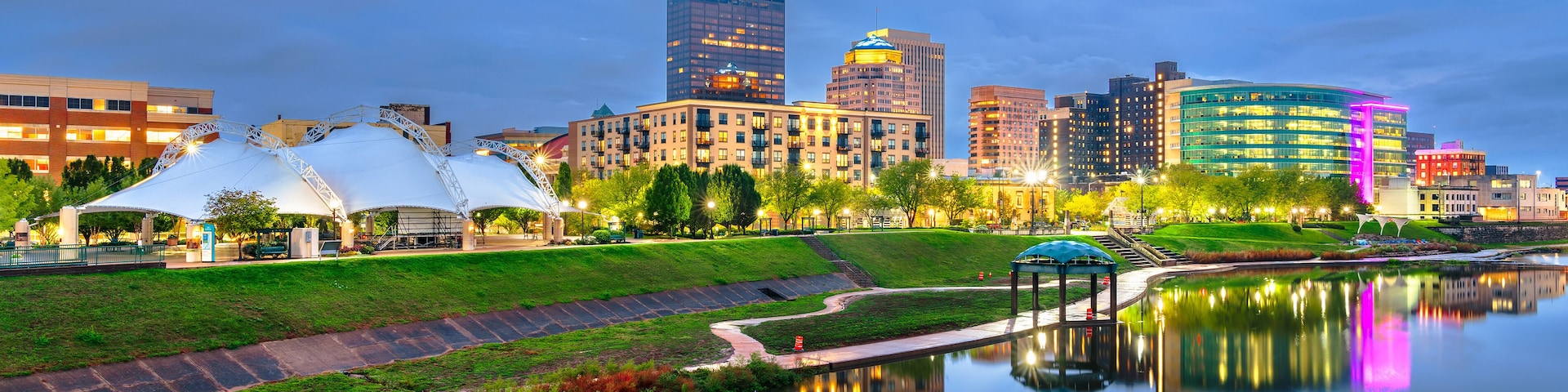 A city skyline at dusk with a calm river reflecting the buildings, a park with a white gazebo, and a modern building with a blue dome.