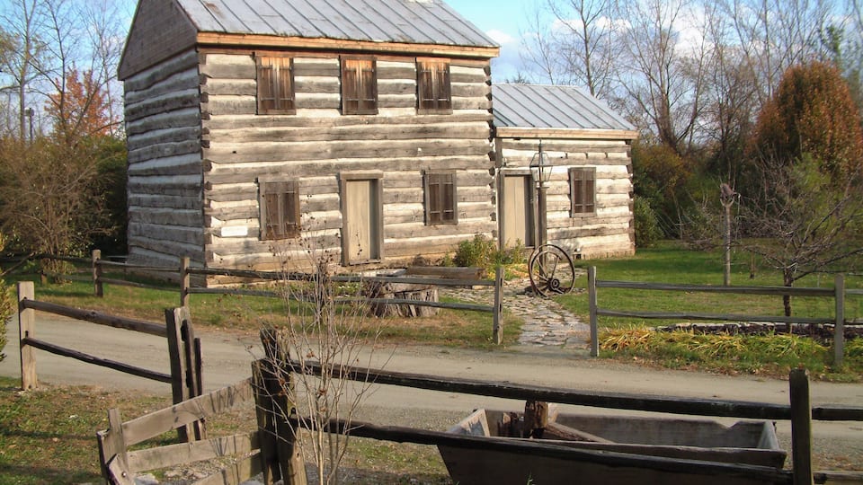 A log cabin with a metal roof and wooden shutters, surrounded by a wooden fence and a stone pathway.