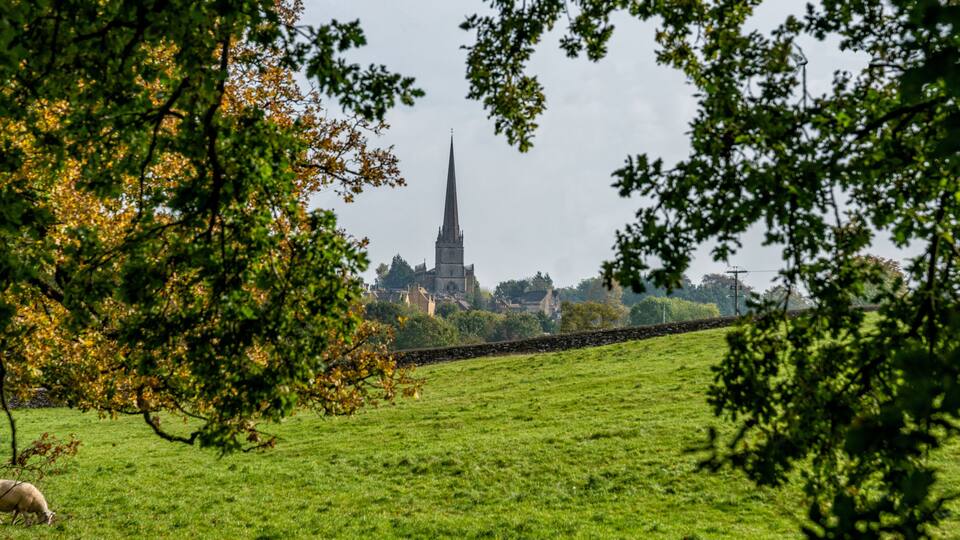 A green field with a stone wall, a sheep, and a church with a tall spire in the background.