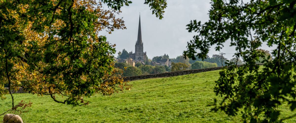 A green field with a stone wall, a sheep, and a church with a tall spire in the background.