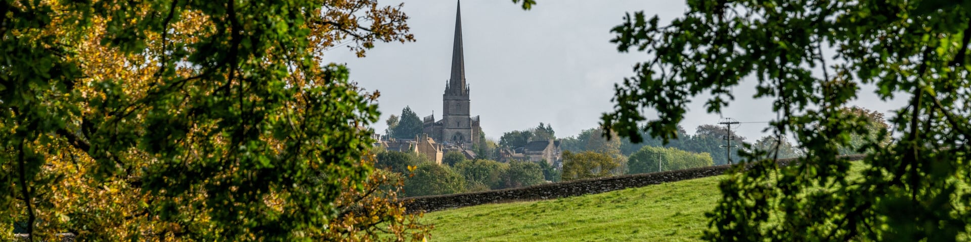 A green field with a stone wall, a sheep, and a church with a tall spire in the background.