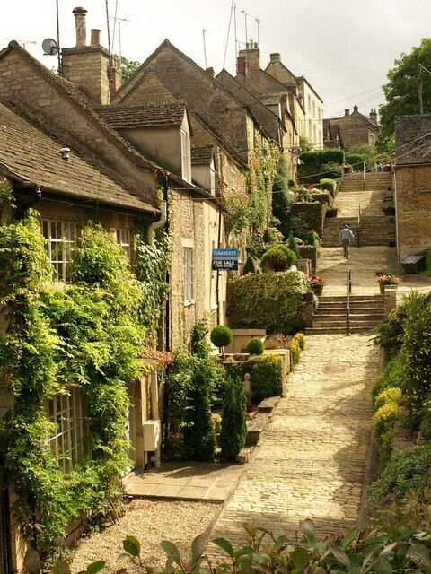 A narrow cobblestone street lined with traditional stone houses, a 'For Sale' sign, and a staircase leading uphill.