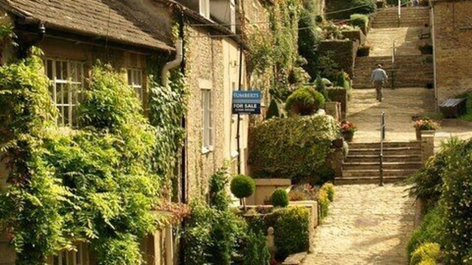 A narrow cobblestone street lined with traditional stone houses, a 'For Sale' sign, and a staircase leading uphill.