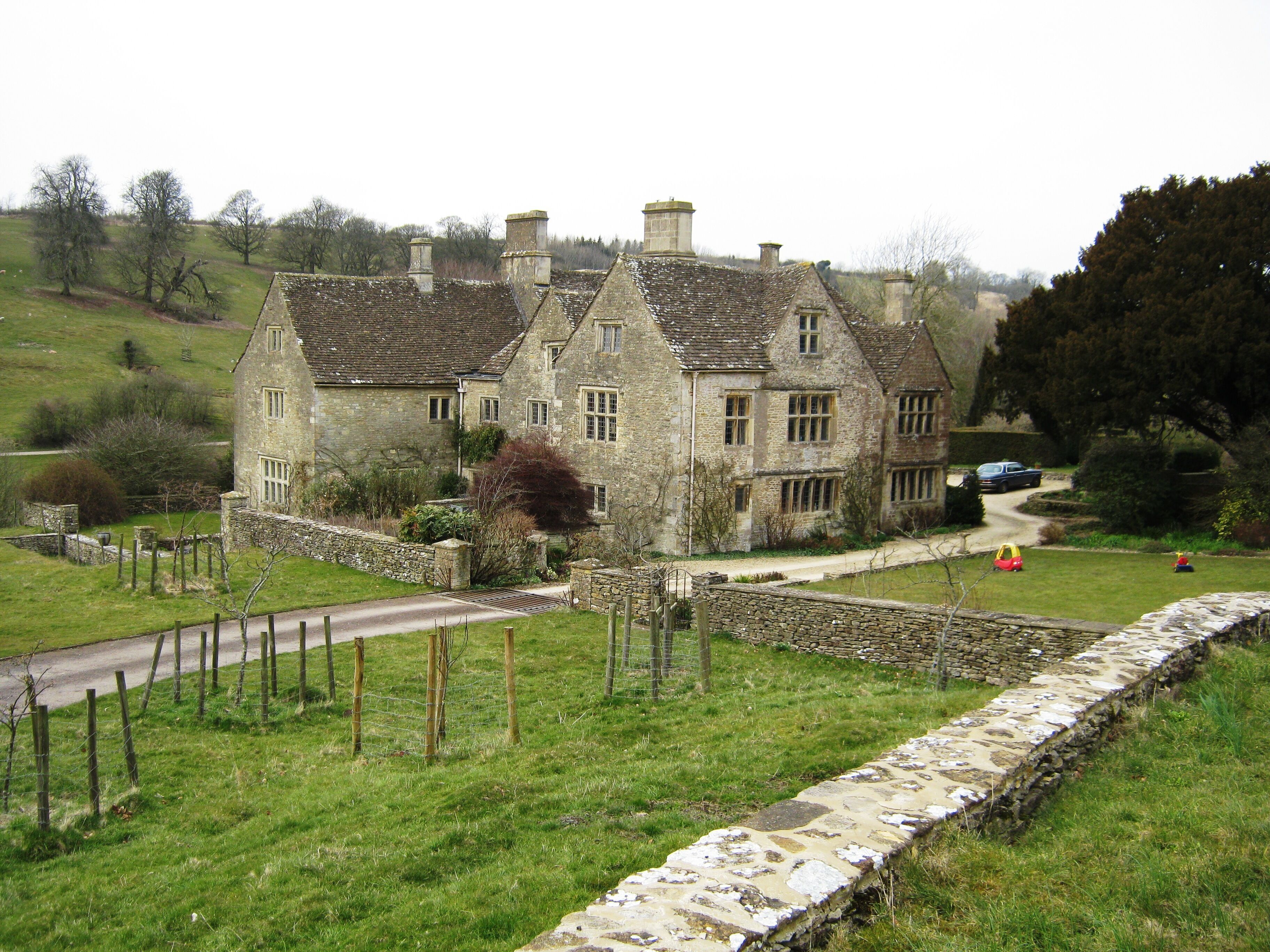A stone house with multiple chimneys, surrounded by a stone wall and a grassy field.