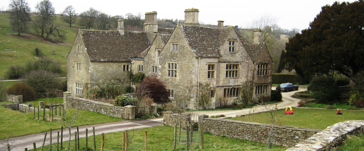 A stone house with multiple chimneys, surrounded by a stone wall and a grassy field.