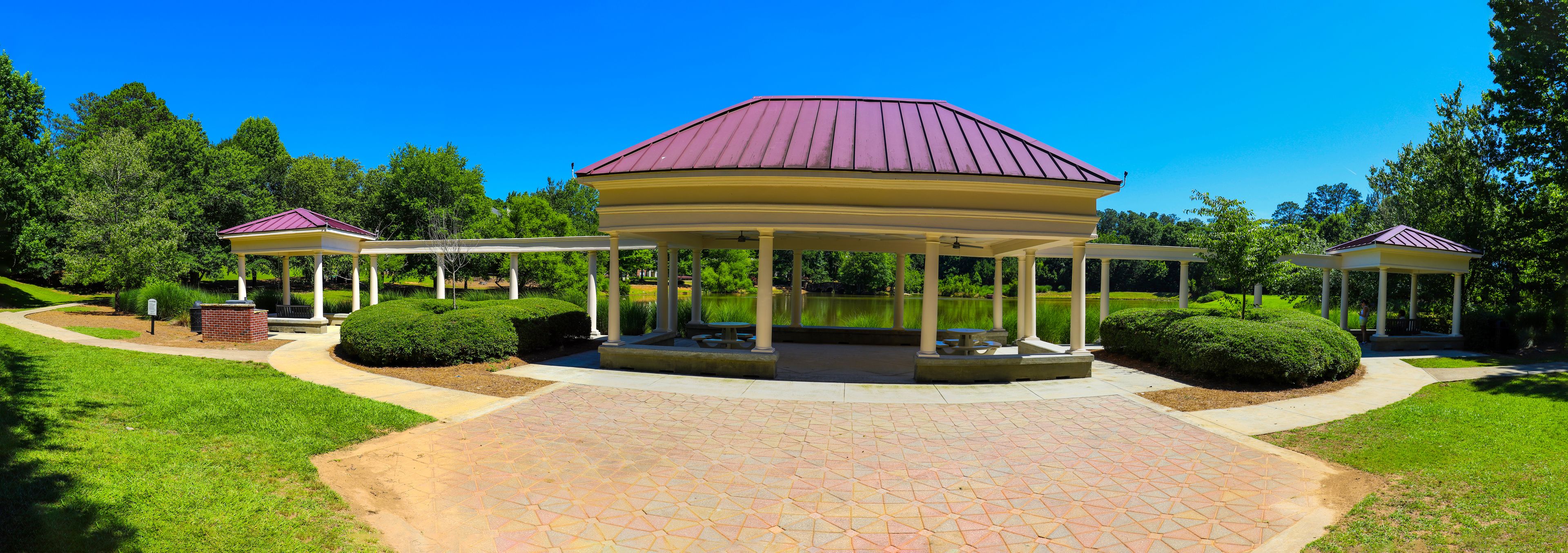 a panoramic shot of a white and red pergola with round pillars on the banks of a silky green pond surrounded by lush green trees, grass and plants with a clear blue sky at Mill Creek Pond