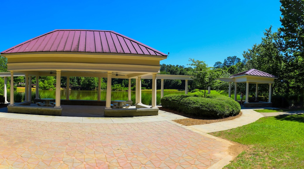 a panoramic shot of a white and red pergola with round pillars on the banks of a silky green pond surrounded by lush green trees, grass and plants with a clear blue sky at Mill Creek Pond
