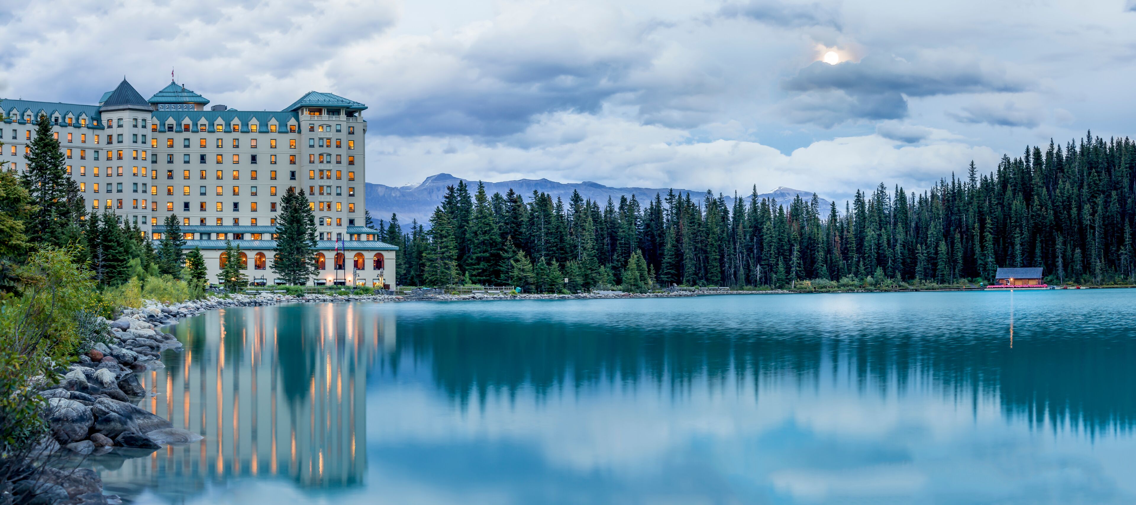 Um hotel grande refletido em um lago calmo, rodeado por floresta densa e montanhas.