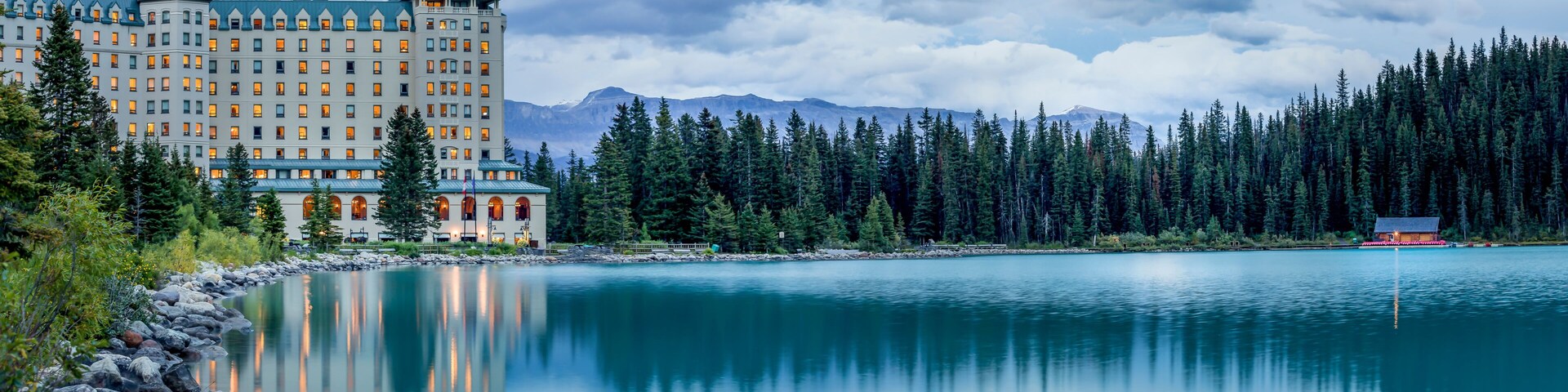 Um hotel grande refletido em um lago calmo, rodeado por floresta densa e montanhas.
