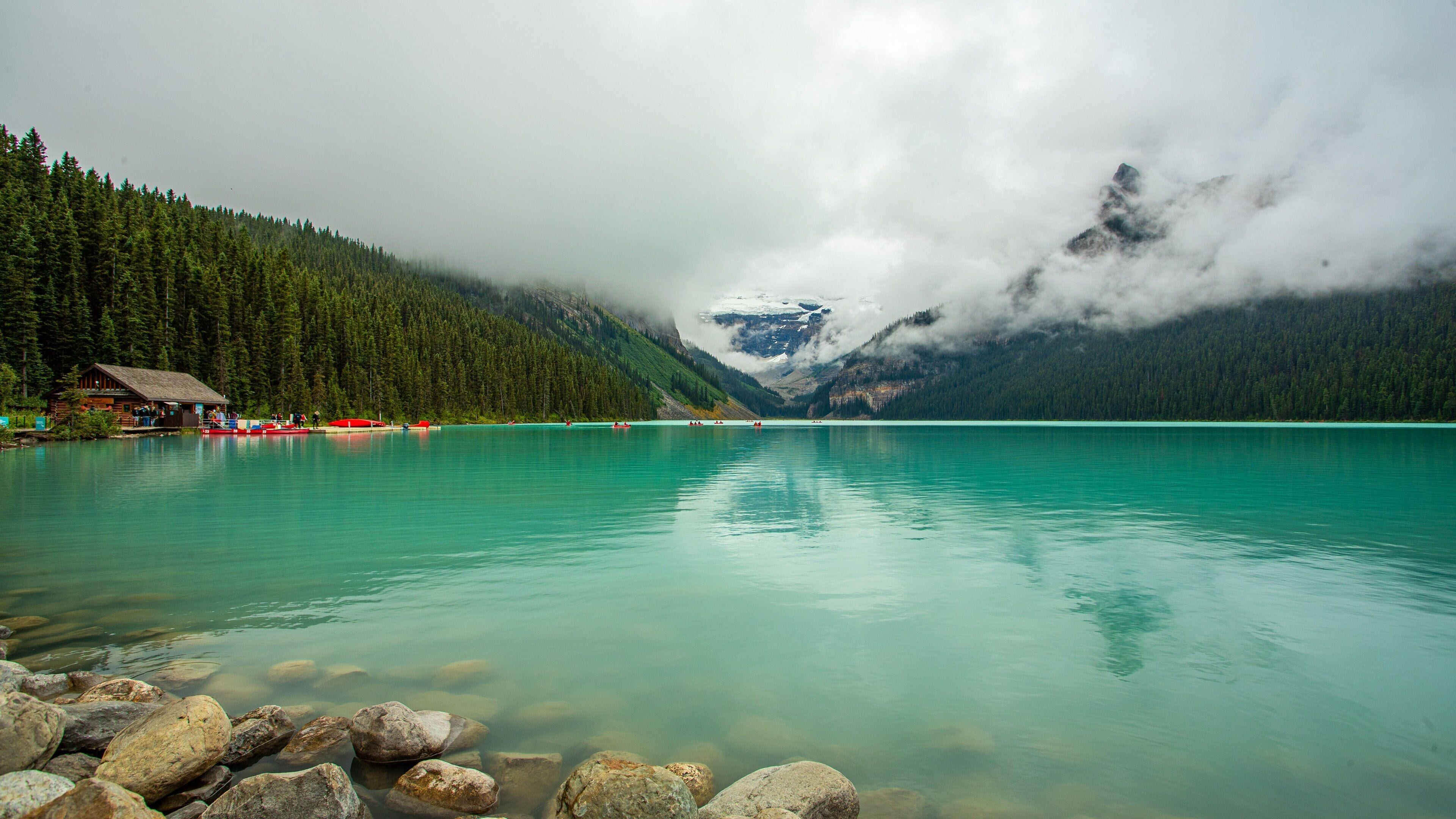 A calm lake with clear turquoise water, surrounded by dense green forests and snow-capped mountains.