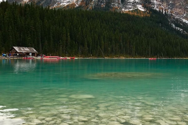 Ein klarer See mit sichtbaren Felsen, eine Hütte und verschneite Berge im Hintergrund.