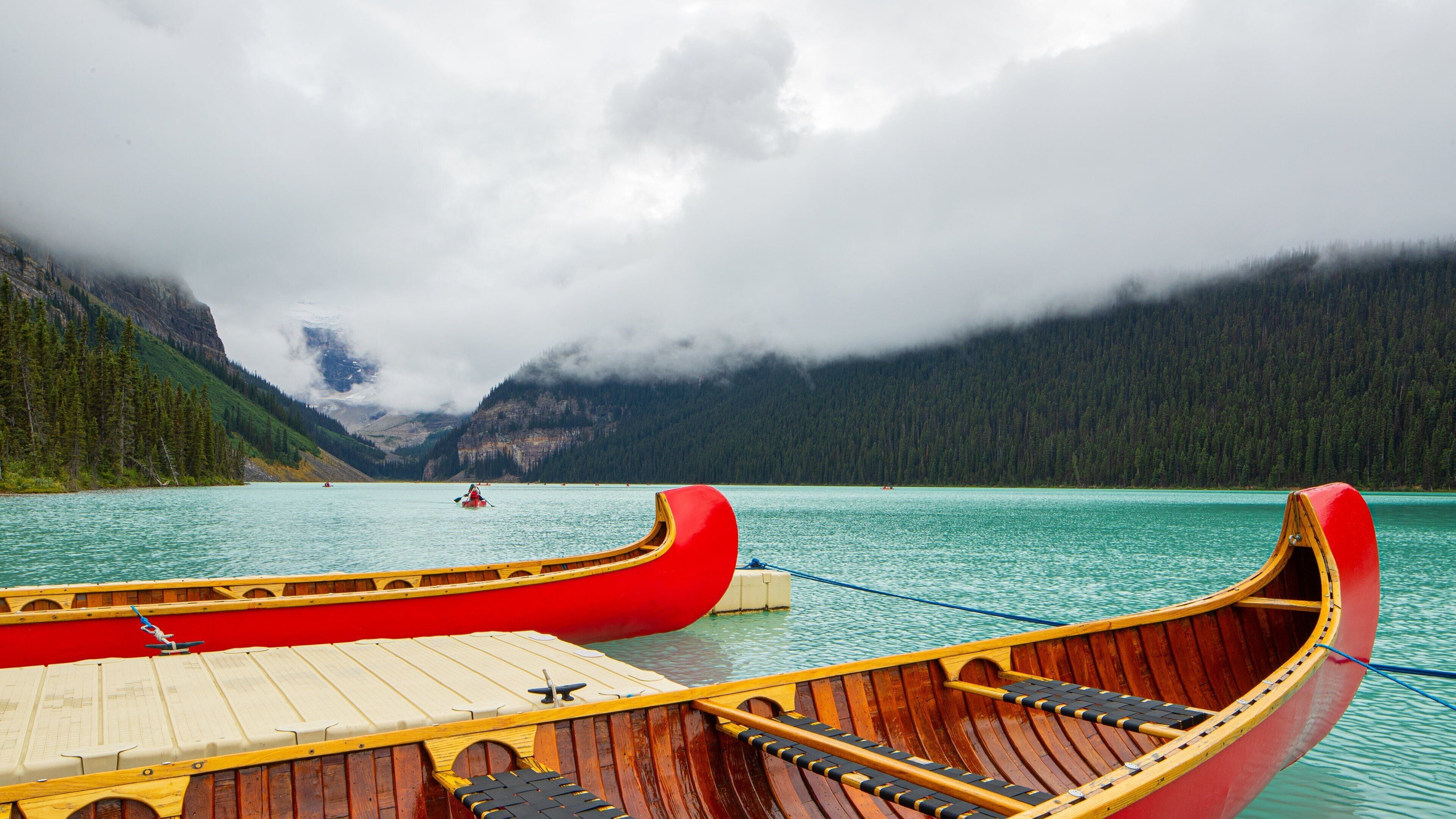 Two red wooden boats on calm turquoise water, with a forested mountain range in the background and a cloudy sky.