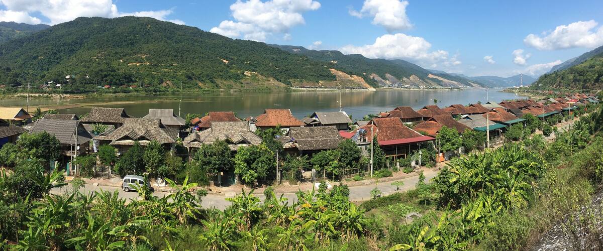 A riverside village with traditional houses, lush greenery, and mountains in the background.
