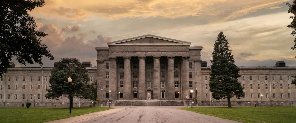 A large neoclassical building with a central pediment and flanking columns, surrounded by a wide, cracked road and green lawns.