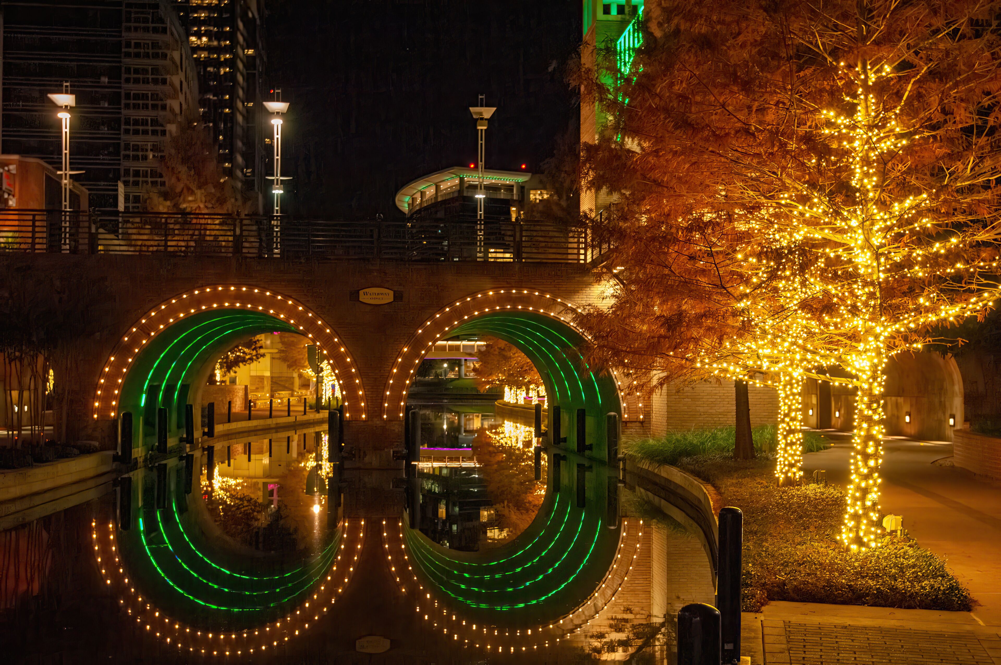 A nighttime urban scene with illuminated green and yellow lights forming circular patterns on a bridge, a tree with yellow lights, and a building with a green-lit corner.
