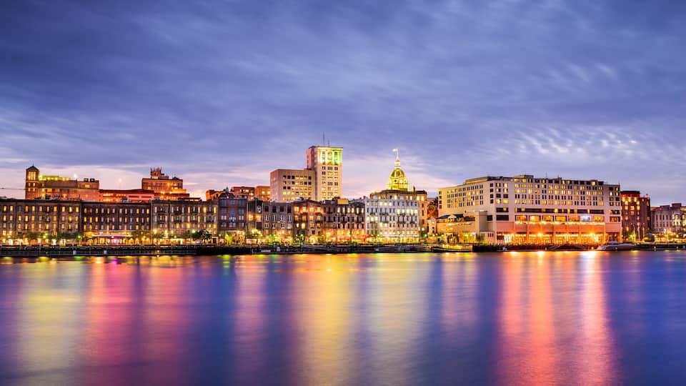A city skyline at dusk with illuminated buildings reflecting on the water.