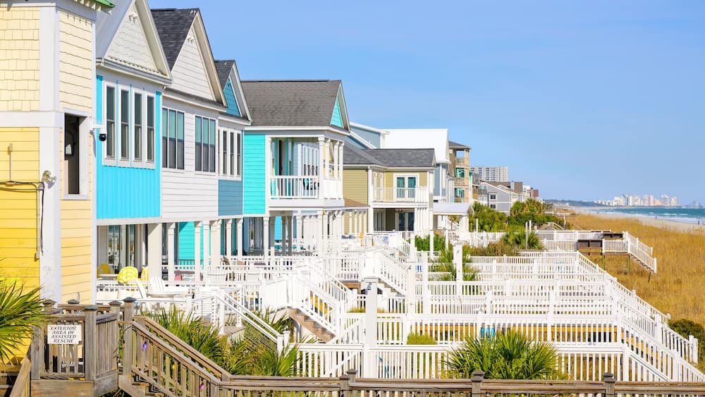 Une rangée de maisons de plage aux couleurs vives, dotées de balustrades blanches et d’un escalier en bois menant à la plage.