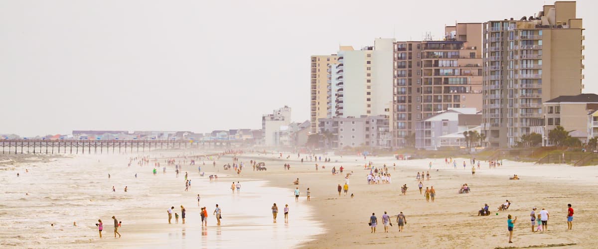 A crowded beach with people walking and swimming, a pier in the background, and tall buildings nearby.