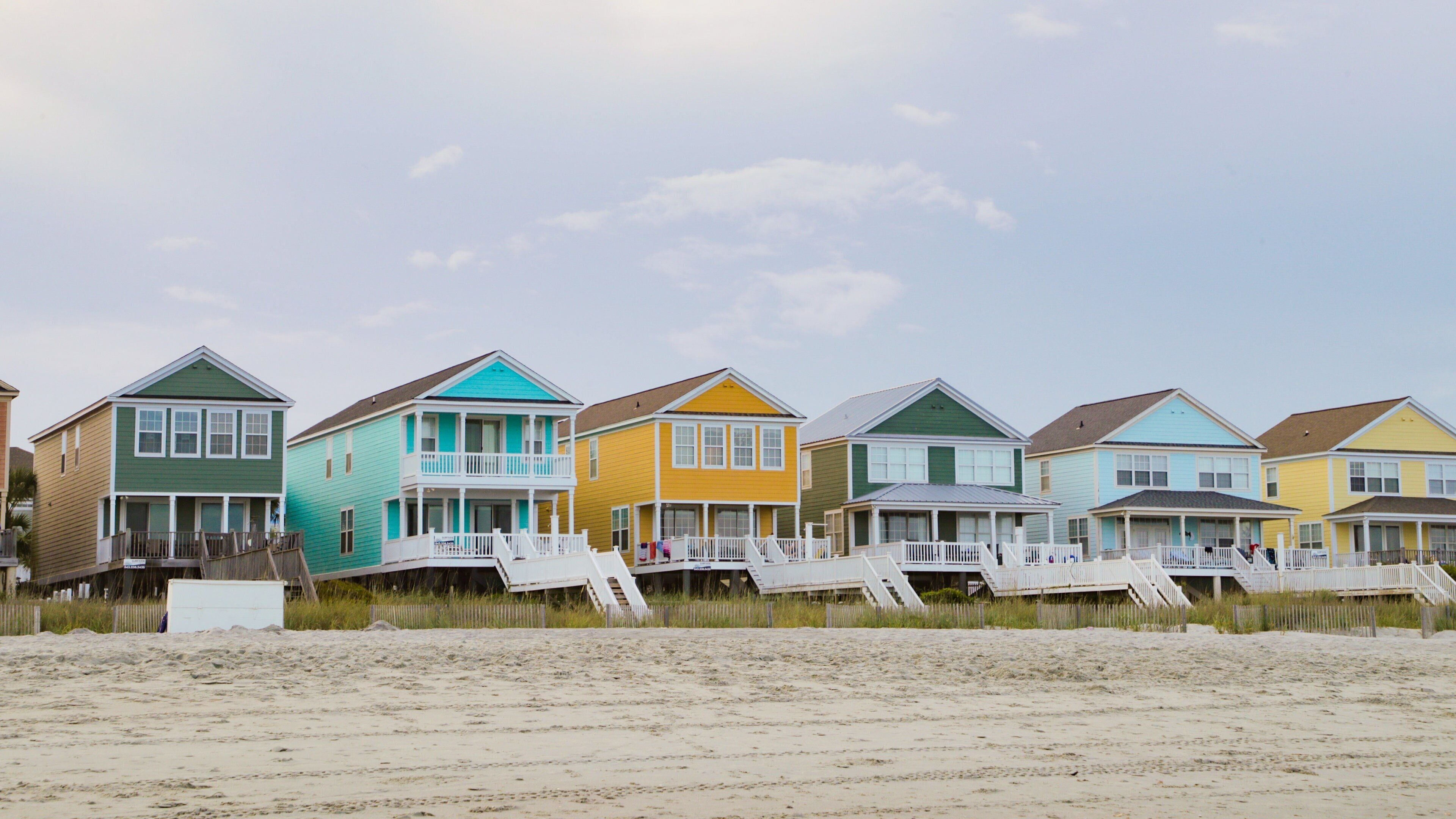 A row of colorful beach houses with white railings and a sandy beach in the foreground.