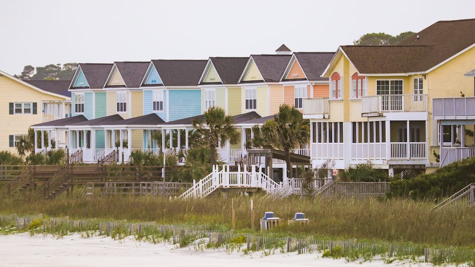 A row of colorful beach houses with white railings and a sandy beach in the foreground.