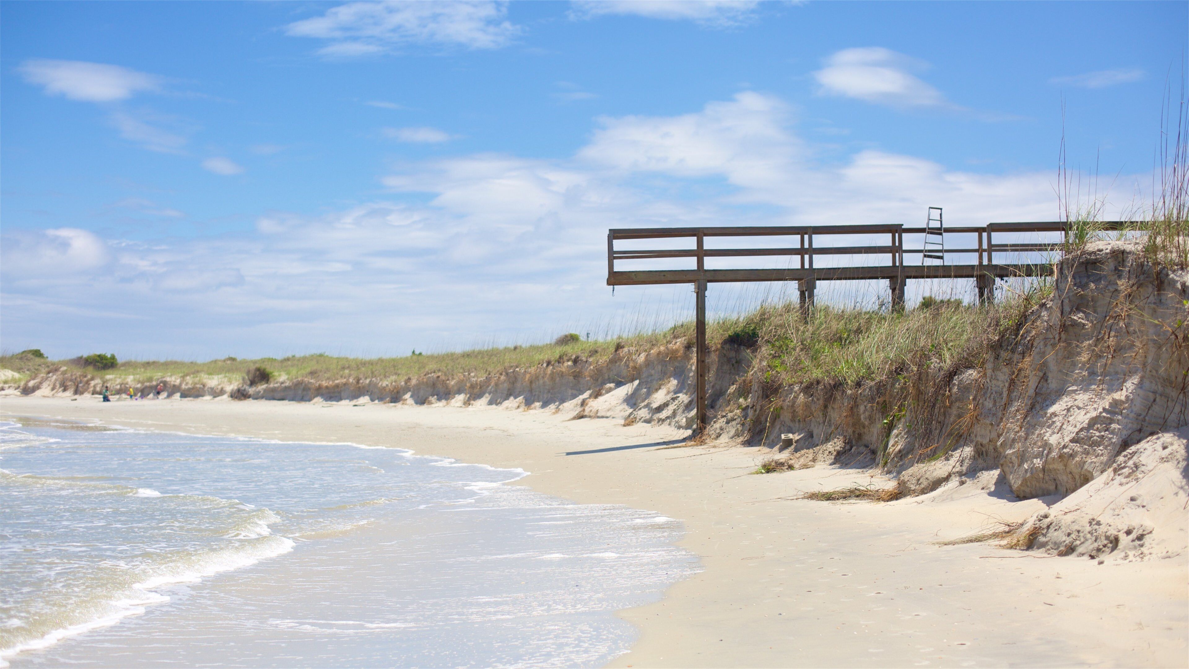 A sandy beach with a wooden structure and a ladder on a grassy cliff.