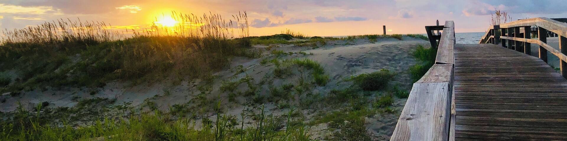 A wooden boardwalk leading to a beach with sand dunes and grasses.
