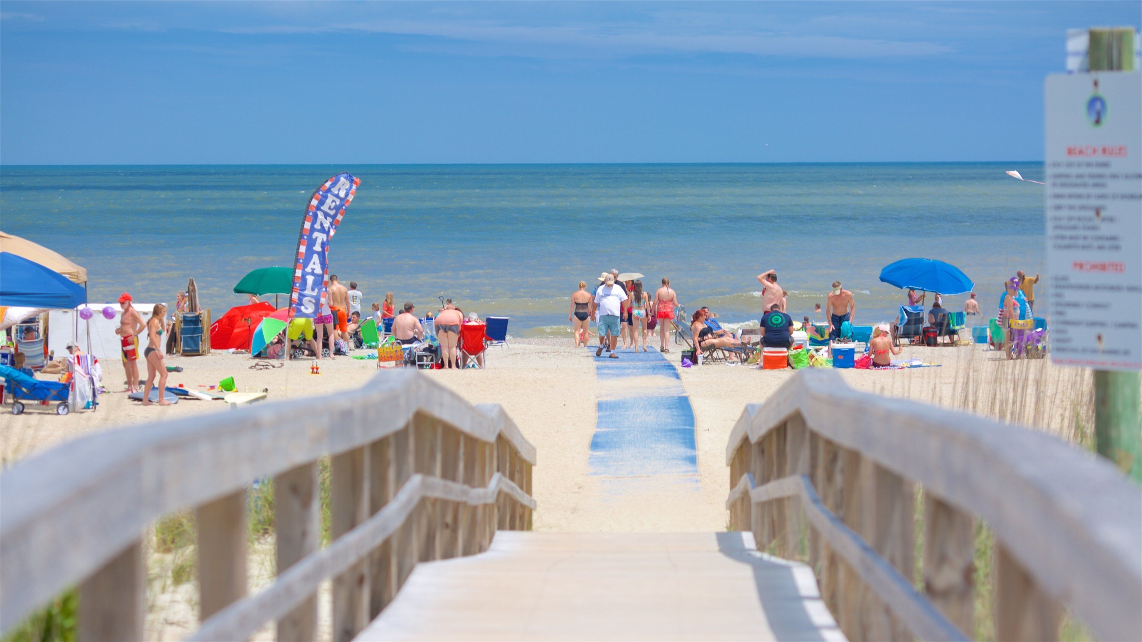 A beach with a wooden boardwalk leading to the water, umbrellas, and people.