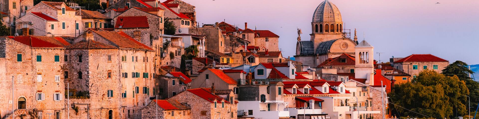 evening at Banj beach with a view of the historic Sibenik city center and fortress under a colorful sunset sky, Croatia.