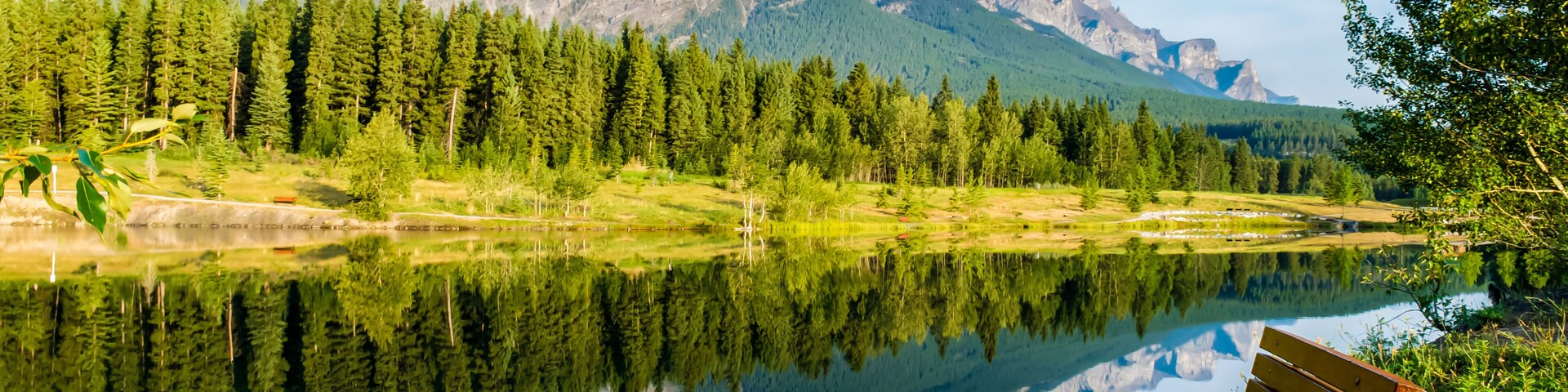 Een serene vijver met de weerspiegeling van een bergketen, omringd door dicht bos en een houten bank.