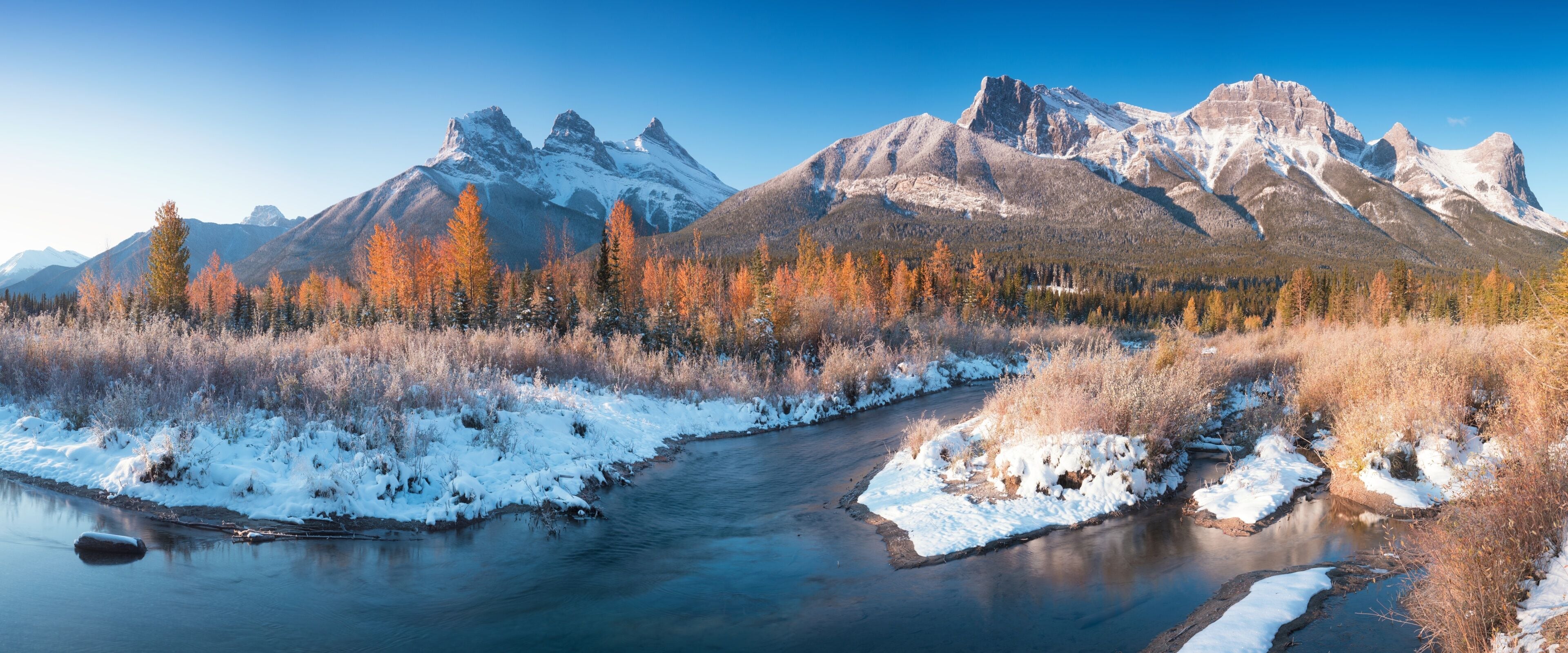 A river flowing through a snowy landscape with autumn-colored trees and snow-capped mountains in the background.