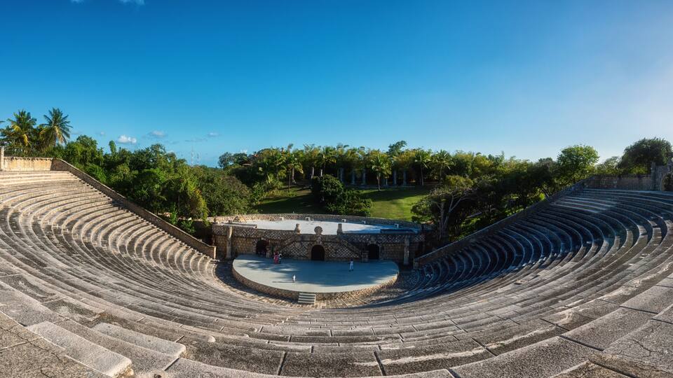 An ancient amphitheater with stone steps and a central stage, surrounded by lush greenery and palm trees.