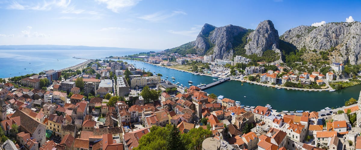 View of terracotta rooftops cascade toward the turquoise river, embraced by towering grey cliffs under a bright blue sky in Omis, Split-Dalmatia County, Croatia.