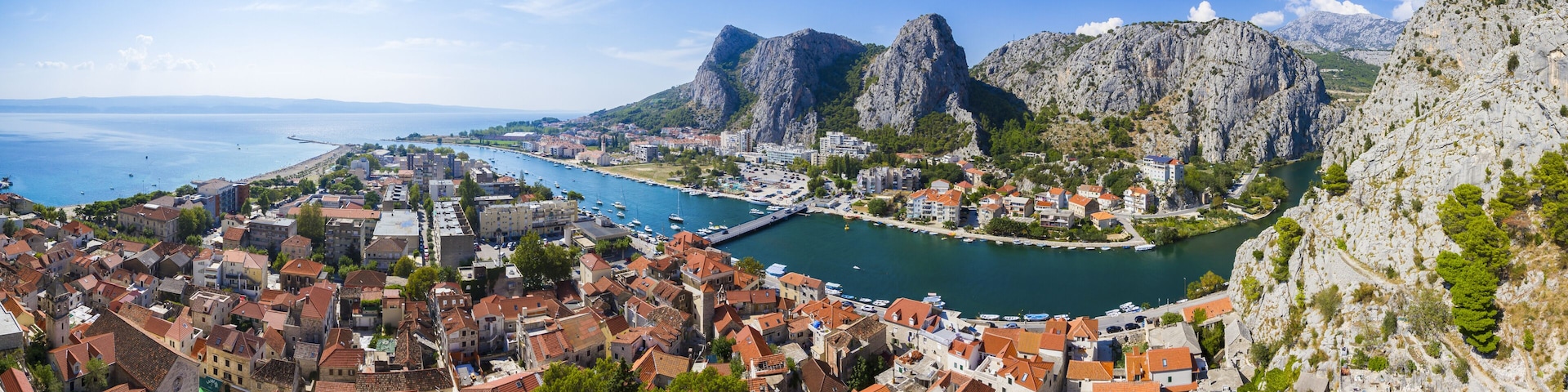 View of terracotta rooftops cascade toward the turquoise river, embraced by towering grey cliffs under a bright blue sky in Omis, Split-Dalmatia County, Croatia.