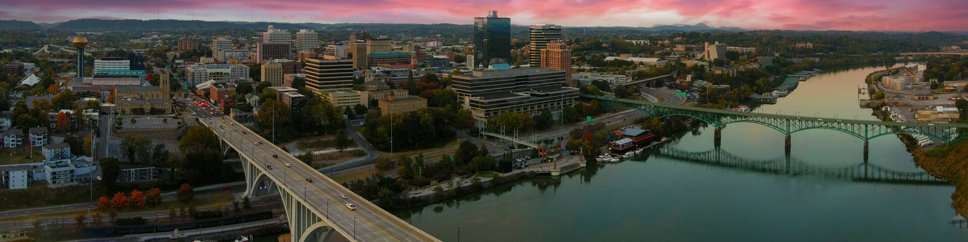 aerial shot of the Henley Street Bridge and Gay Street Bridge over the Tennessee River surrounded by autumn colored trees, lush green trees and office buildings with powerful clouds at sunset