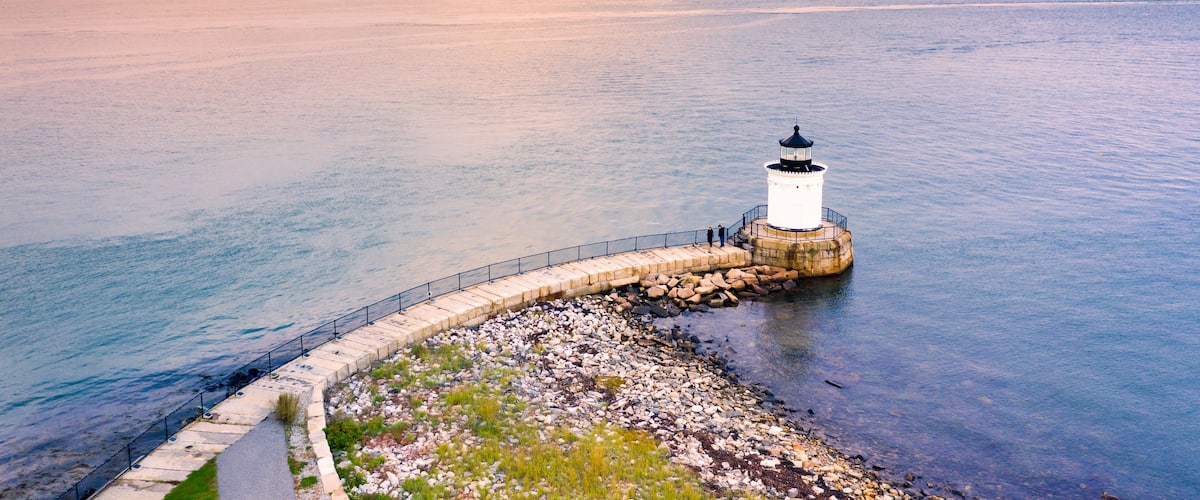A lighthouse on a rocky breakwater, surrounded by calm water and distant land.