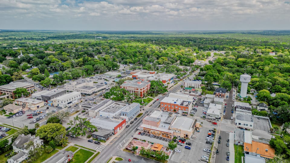 An aerial view of a town with a mix of commercial and residential buildings, surrounded by green landscapes.