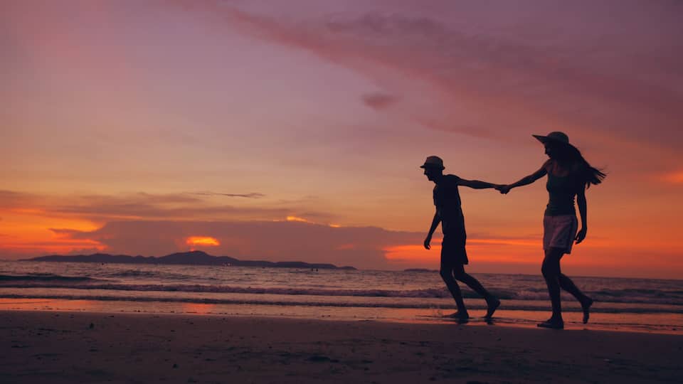 A sunset over a beach with silhouettes of two people holding hands.