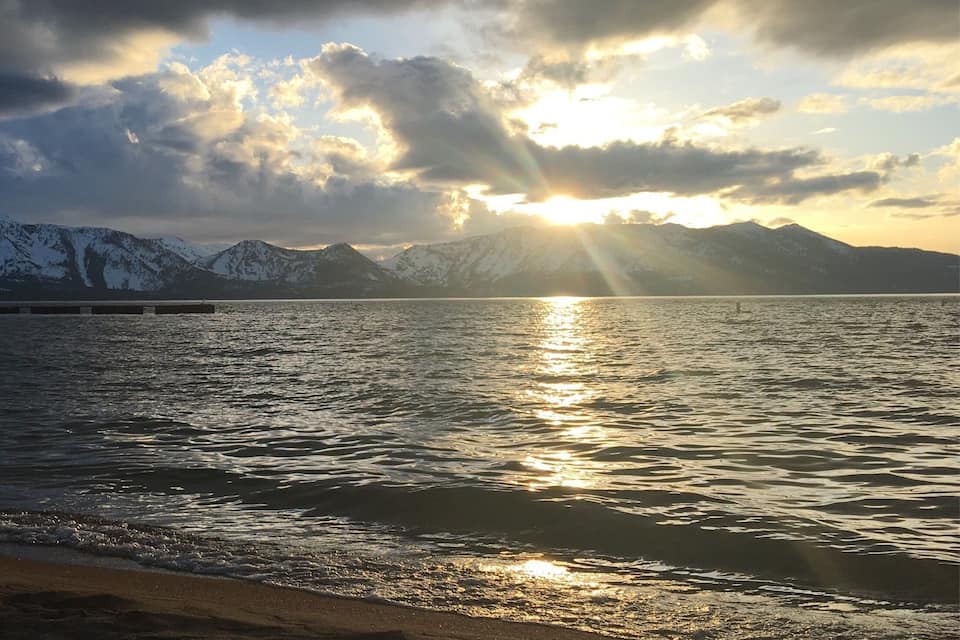 Uma praia com uma cadeia de montanhas ao fundo e o sol se pondo sobre a água.