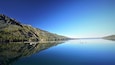 Ein ruhiger See mit einem Steg, umgeben von bewaldeten Bergen unter blauem Himmel.