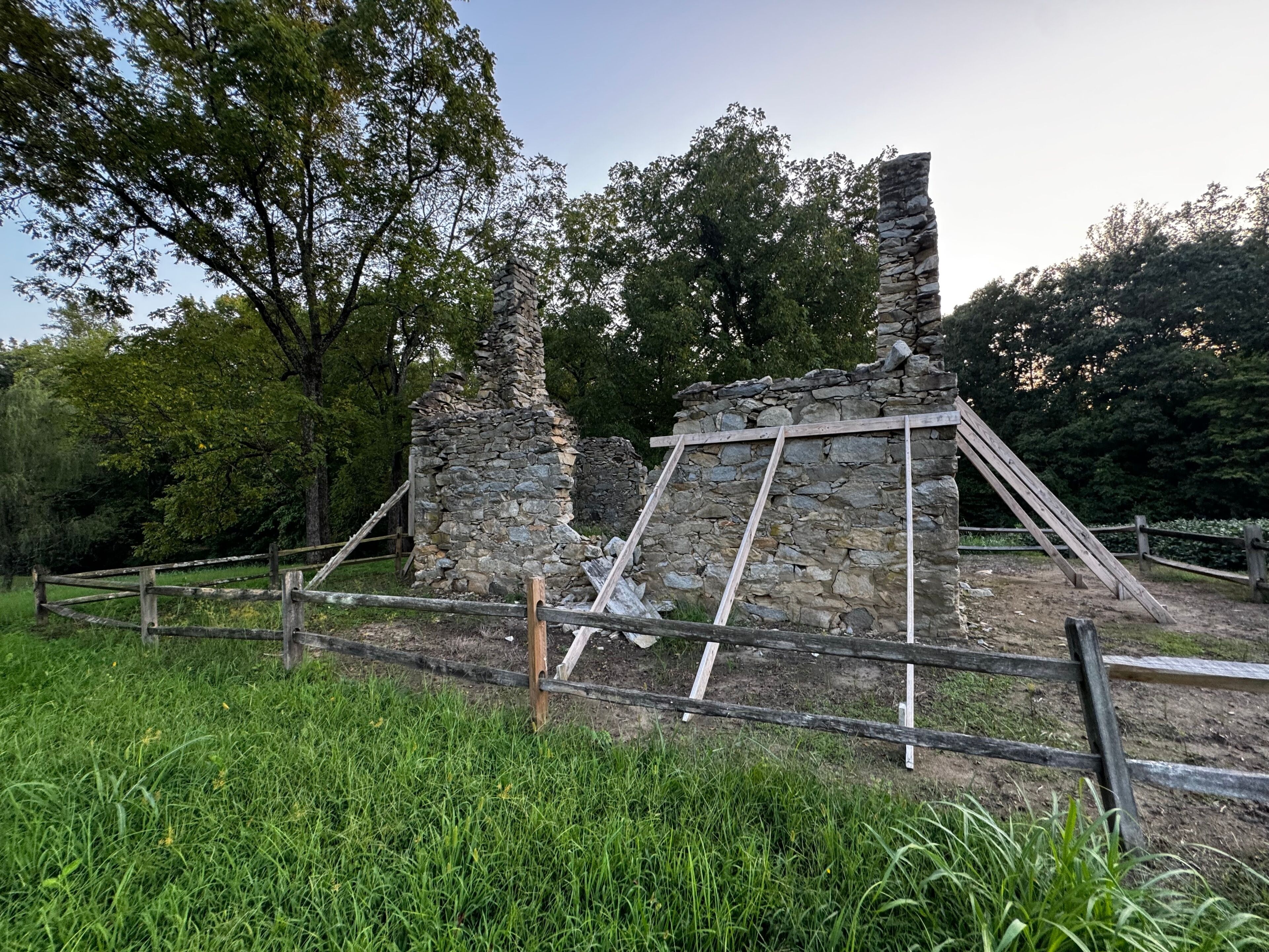 A stone ruin with wooden scaffolding, surrounded by a wooden fence and lush greenery.