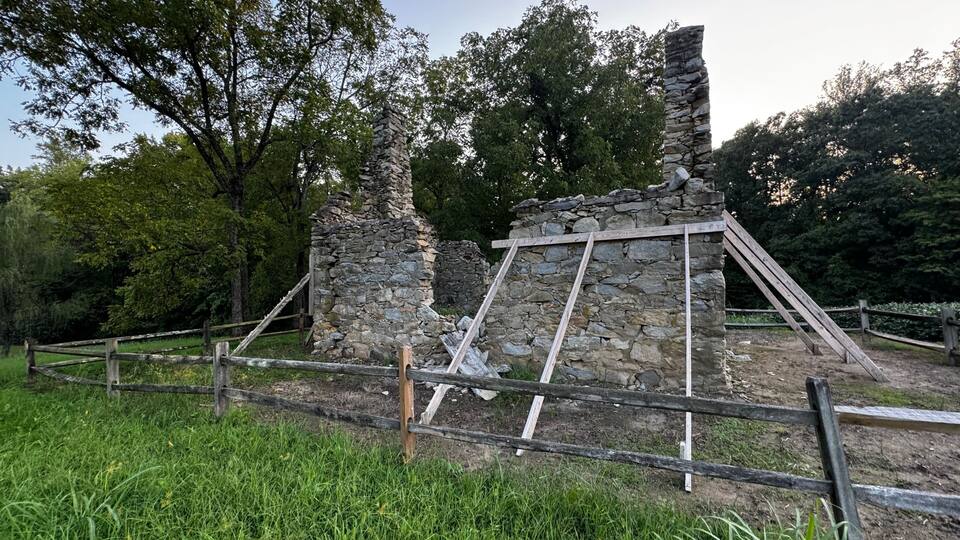 A stone ruin with wooden scaffolding, surrounded by a wooden fence and lush greenery.