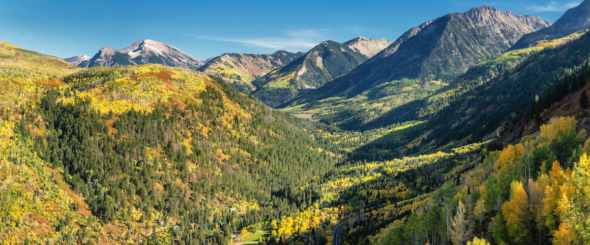 A mountainous valley with autumn foliage, a winding road, and distant peaks.