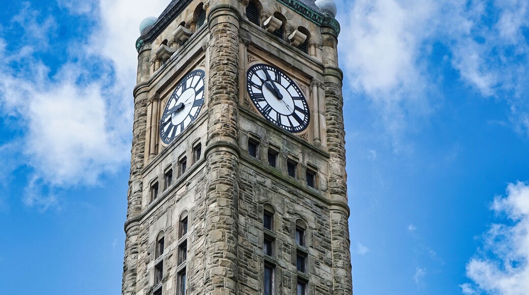 Una alta torre de piedra con un campanario que muestra dos relojes, sobre un cielo azul con nubes.