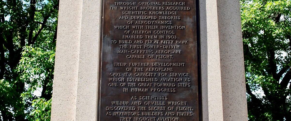 Una placa de bronce montada sobre un pedestal de piedra con la figura alada de un ángel, rodeada de frondosos árboles verdes.
