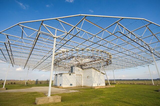 A large metal structure with a roof, possibly a greenhouse or agricultural facility, surrounded by grass and fields.